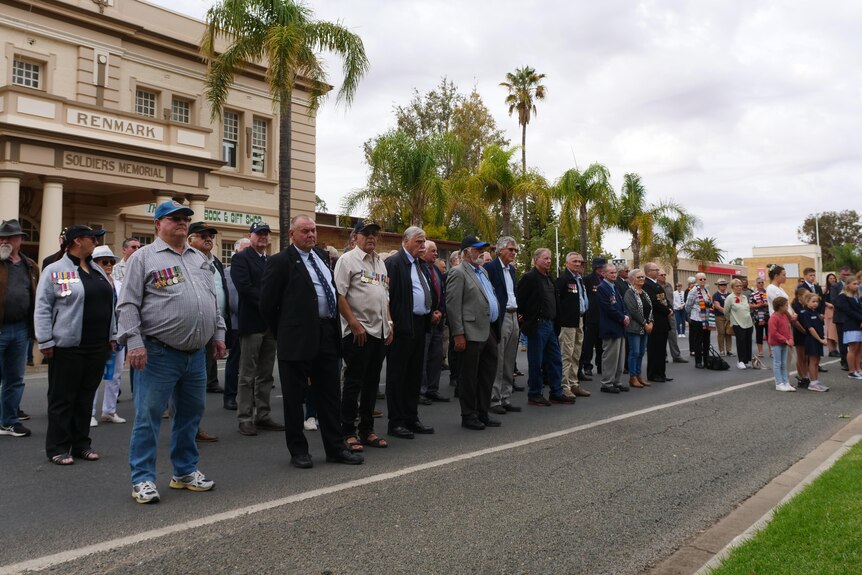A group of 50 people stand under a cloudy sky in front of a building that says renmark soldiers memorial