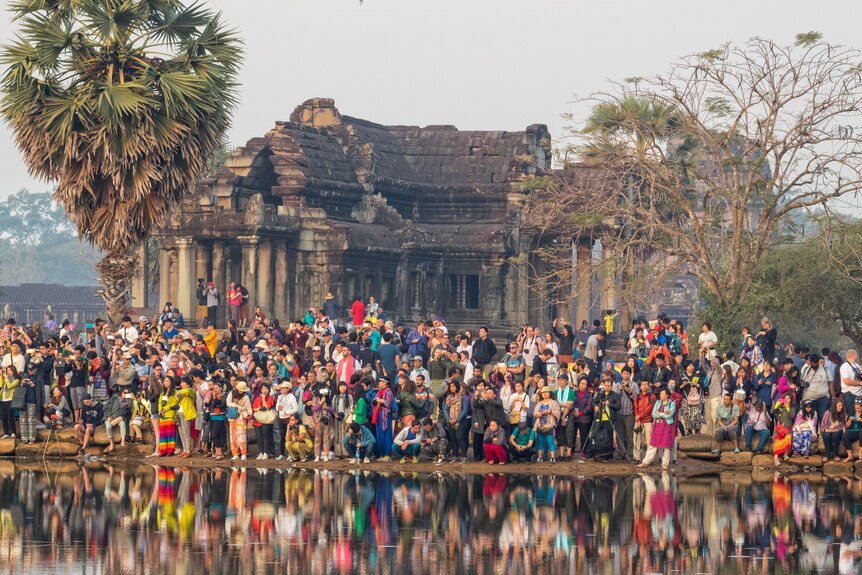 A large group of people stand in front of a temple and a lake