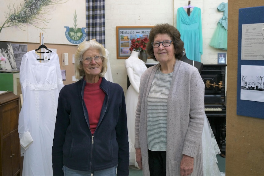 Two women stand smiling at the camera, surrounded by wedding dresses and informative posters that are part of a museum display. 