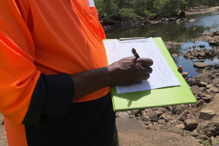A man in a high-vis shirt writing on a green clipboard with a creek behind