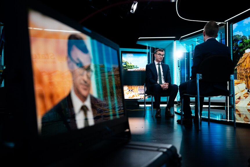 John Carey sits with a serious contemplative expression in a TV studio. His face is broadcast on a monitor in the foreground.