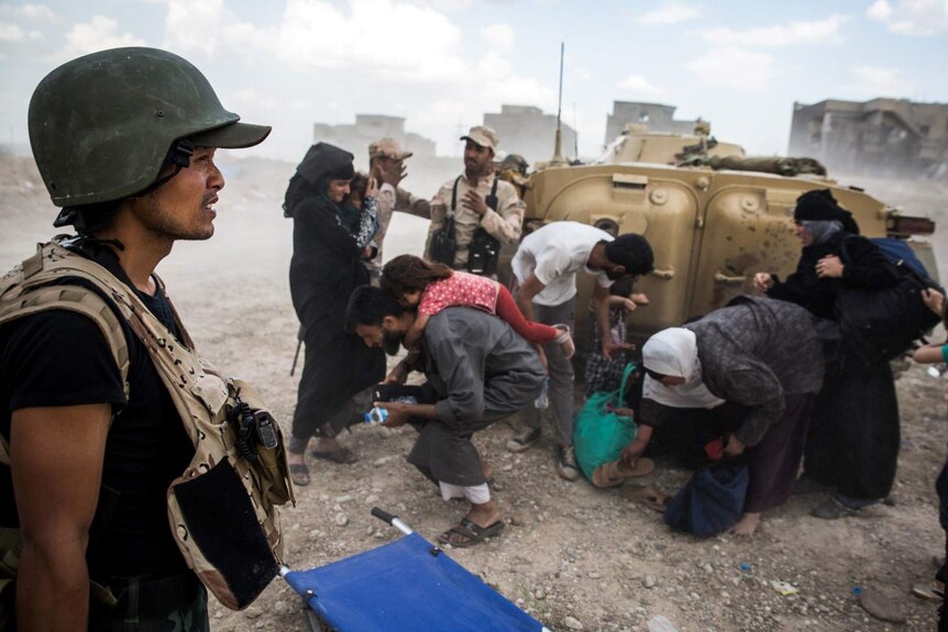 A Burmese man in military armour squints through dust as Iraqi families crouch behind an armoured vehicle.