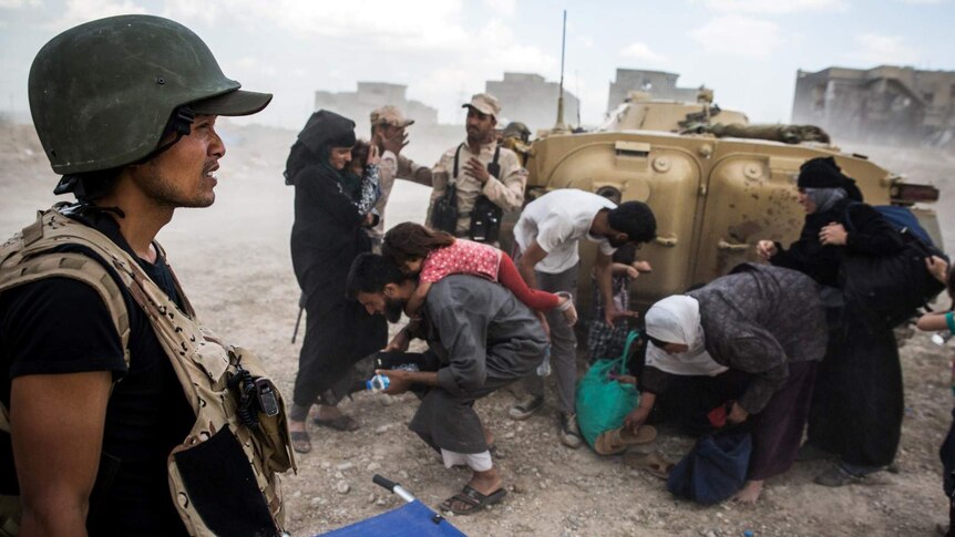 A Burmese man in military armour squints through dust as Iraqi families crouch behind an armoured vehicle.