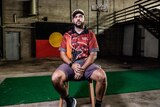 A man sits in a chair in a run down building with an aboriginal flag behind him. 