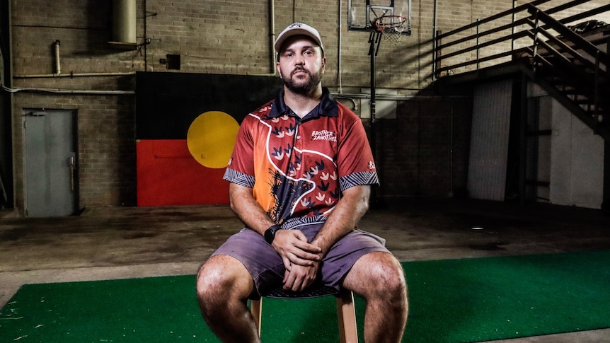 A man sits in a chair in a run down building with an aboriginal flag behind him. 