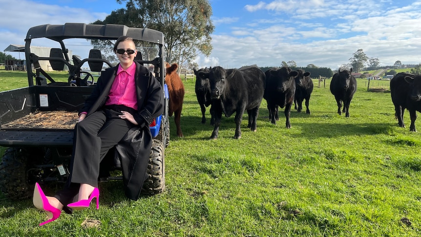 Brodie Pyle in a pink blouse and black blazer and sunglasses sitting on a quad bike next to black cows looking at the camera