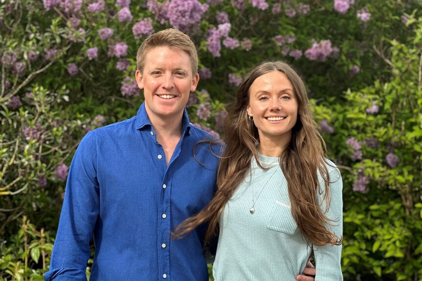 Hugh Grosvenor and Olivia Henson wearing casual clothes pose in front of a garden of flowers on a sunny day.