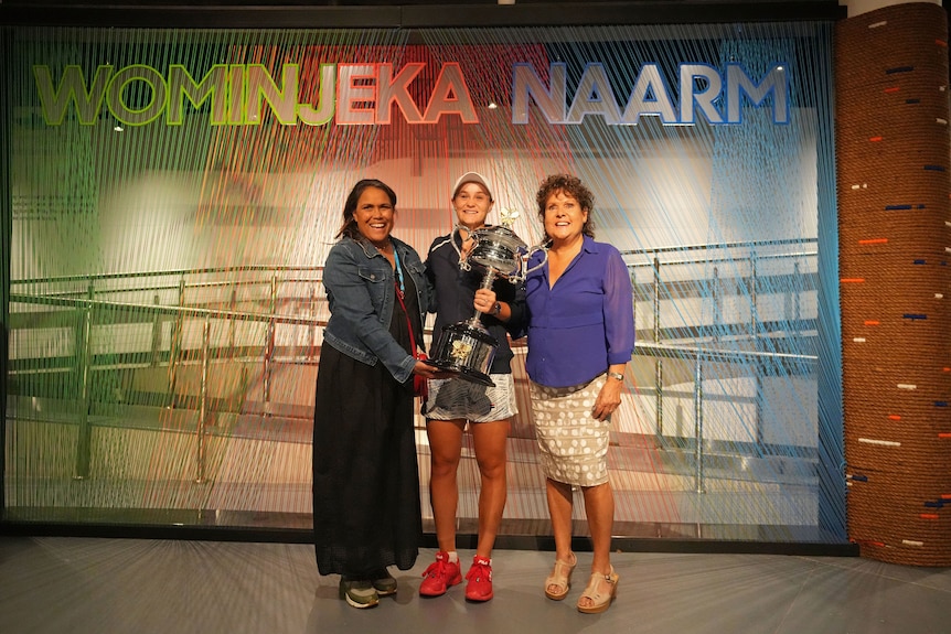Three women stand in a row, with the middle woman holding her tennis trophy