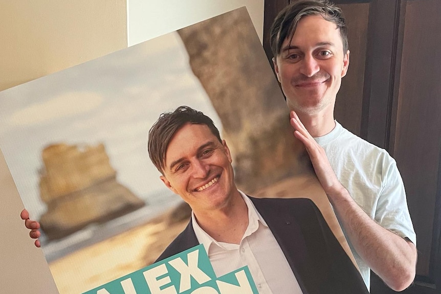 a man holds an election campaign poster of himself and smiles at the camera