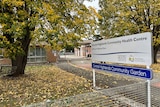 A sign sits outside a building in front of a tree with yellow autumn leaves.