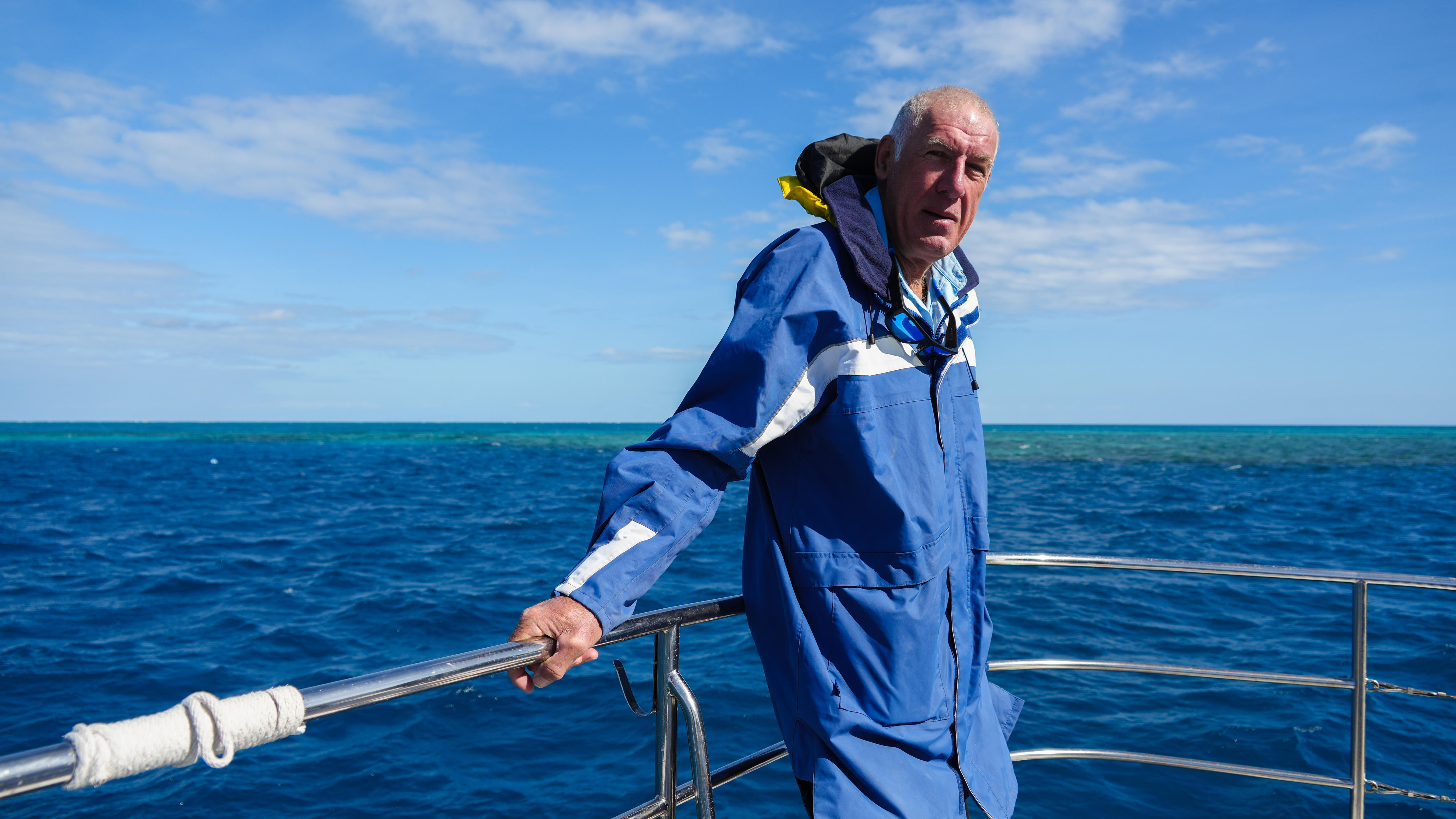 A man standing on the front of a boat holding onto the hand rail with water behind.