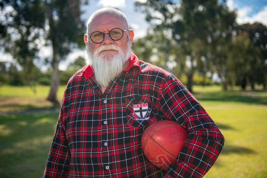 A man holds a football under his arm at a park and looks at the camera.