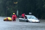Two men wearing helmets and lifejackets push a dinghy next to a car in waist-high water.