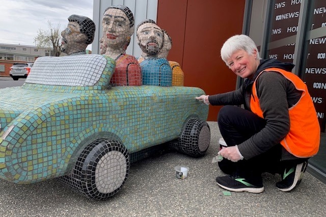A woman in high vis crouching down next to colourful sculpture of a car with figures in it