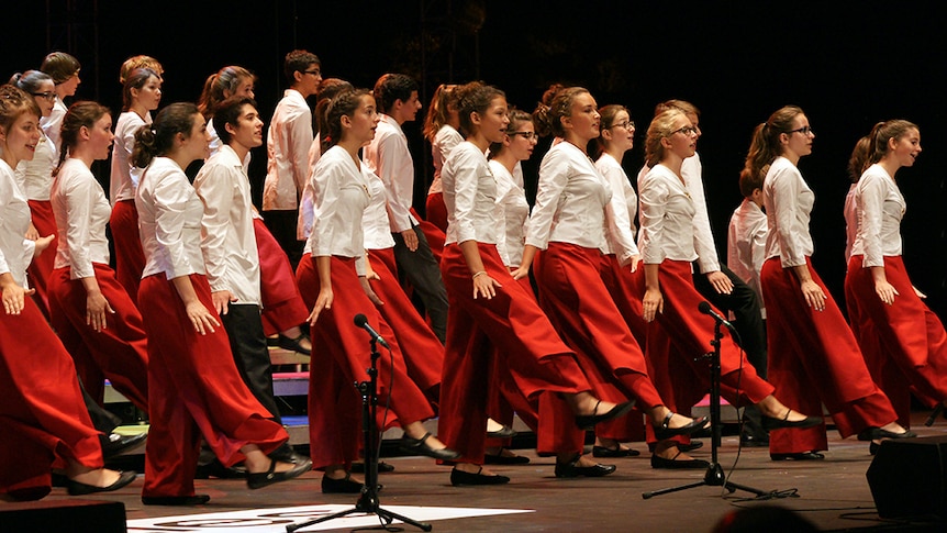 A choir of young students sing and dance wearing white shirts and red or black pants.