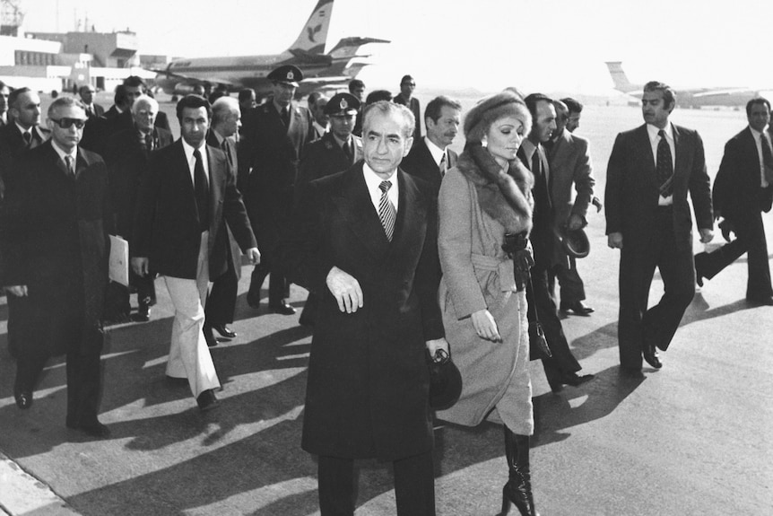 The Shah and his wife walk on tarmac with crowd behind them.
