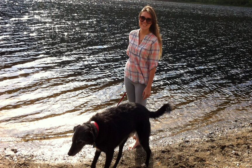A young woman with a dog on a lead smiling at the camera on the shores of a lake.
