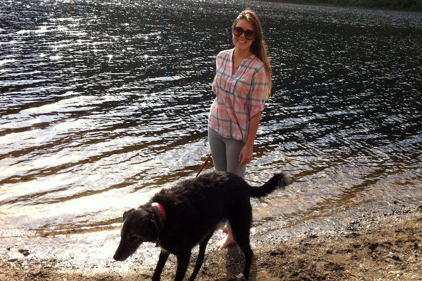 A young woman with a dog on a lead smiling at the camera on the shores of a lake.