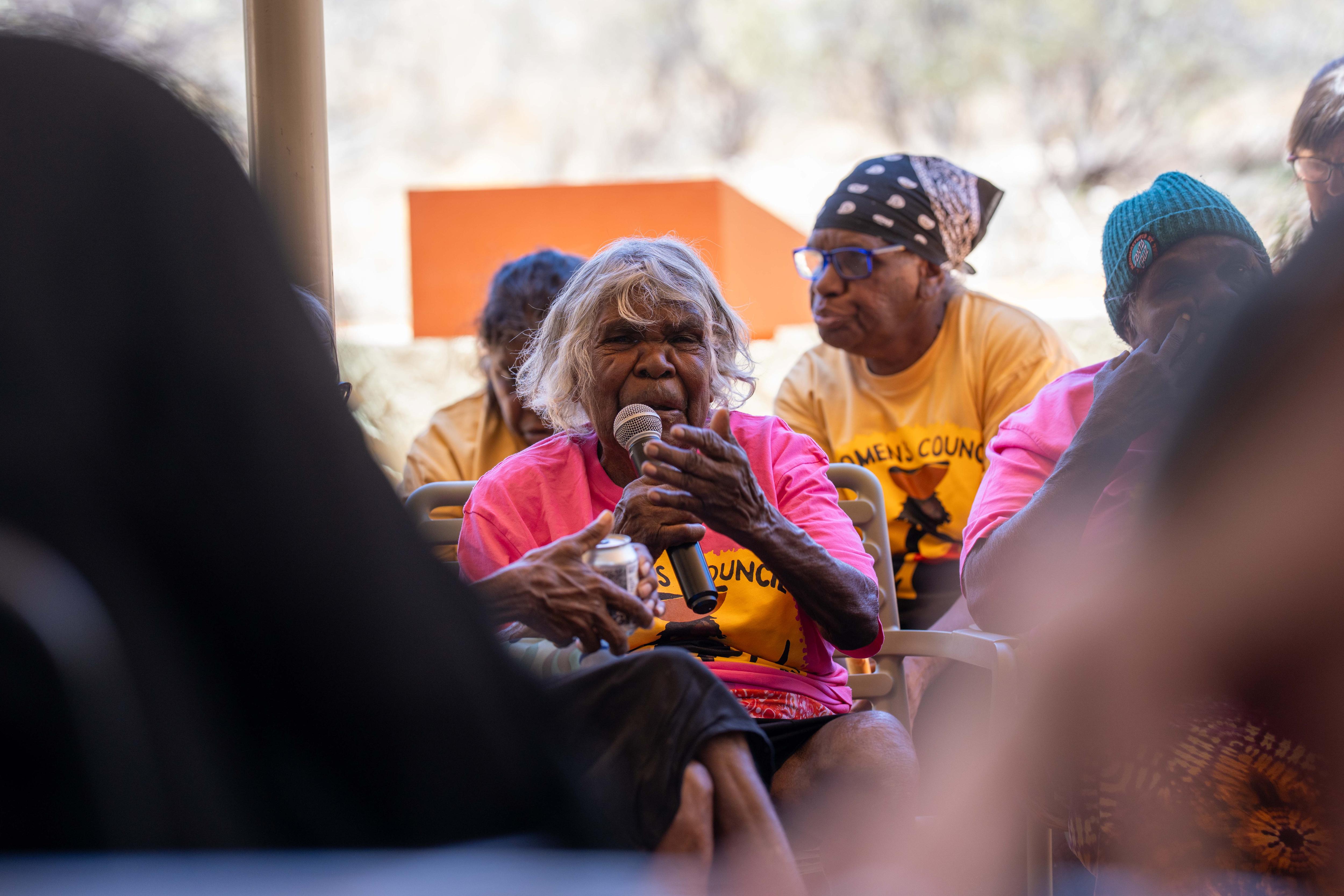 A seated Aboriginal woman holds a microphone and gestures with her free hand.