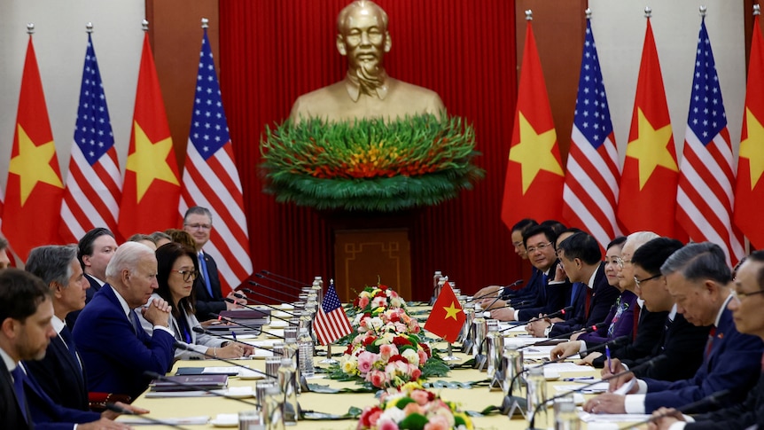 US and Vietnamese officials sit on either side of a long table with a statue of Ho Chi Minh in the background.