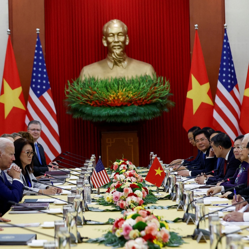 US and Vietnamese officials sit on either side of a long table with a statue of Ho Chi Minh in the background.