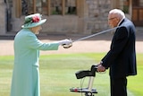 The Queen places a sword on the left shoulder of Sir Tom Moore who is wearing a suit and leaning on a walker