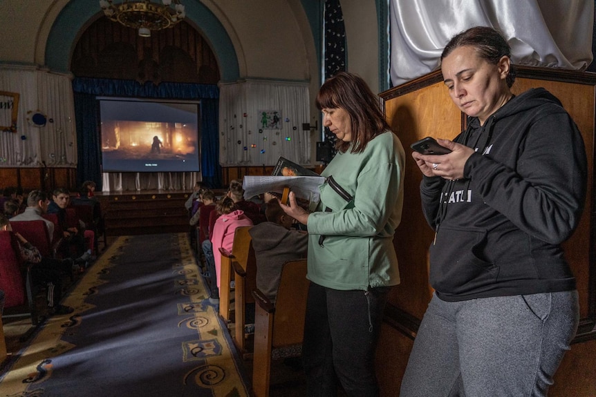 Two women look on their phones in Lviv while standing in a hall.