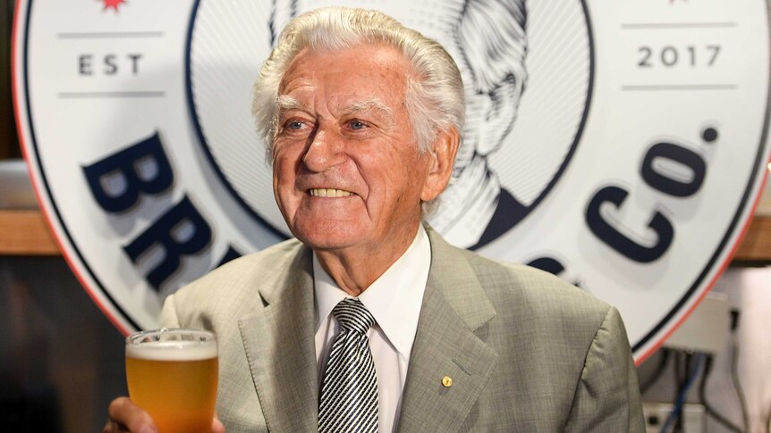 Bob Hawke wears a grey blazer and looks to the left as he holds a pint. His hair's grey, he's smiling and a logo's behind.