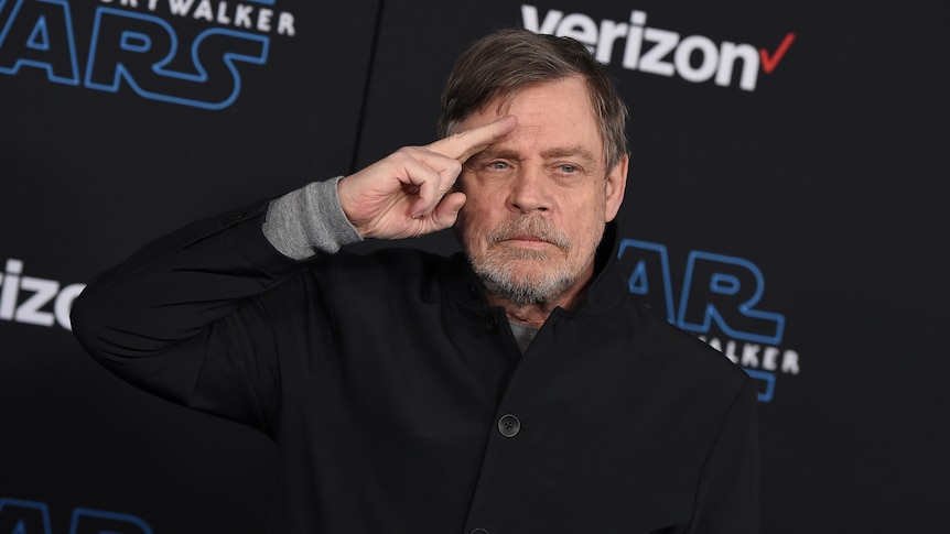 An older man with grey hair and a short beard salutes with two fingers in front of a black Star Wars backdrop.