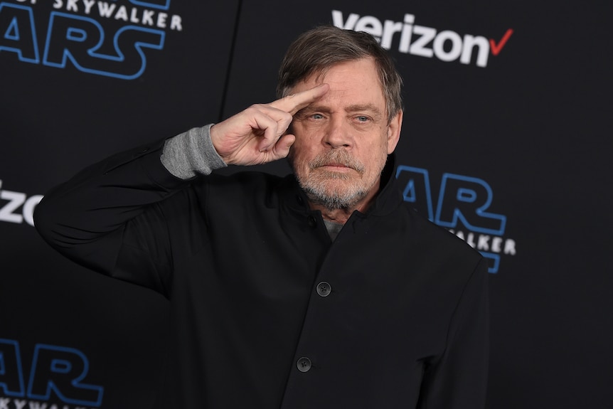 An older man with grey hair and a short beard salutes with two fingers in front of a black Star Wars backdrop.