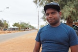 A man wearing a hat smiles with a road street stretching into the distance and a camp dog sulking in the bottom right corner