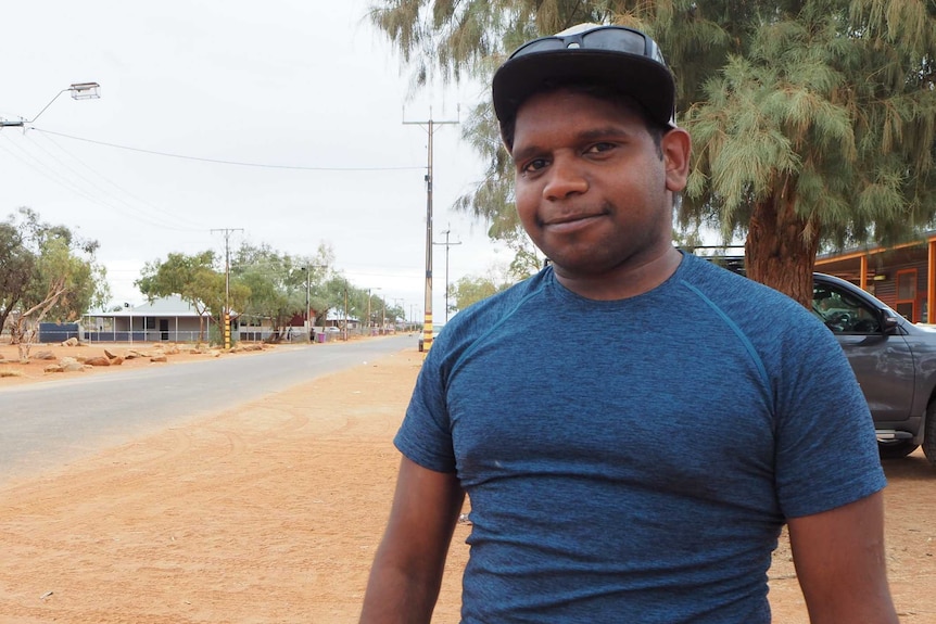 A man wearing a hat smiles with a road street stretching into the distance and a camp dog sulking in the bottom right corner