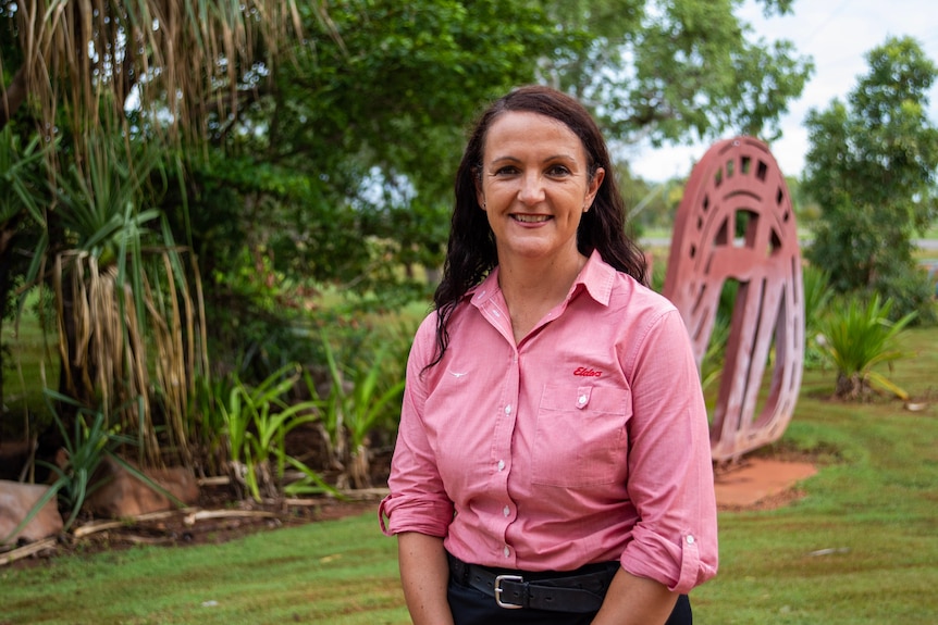 A long-haired woman, wearing a pink shirt and black skirt, stands in front of greenery on a dreary day in Katherine.