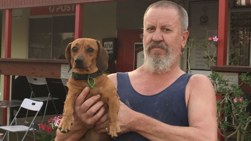 A man with a grey beard, wearing a blue singlet, holds a tan miniature dachshund.