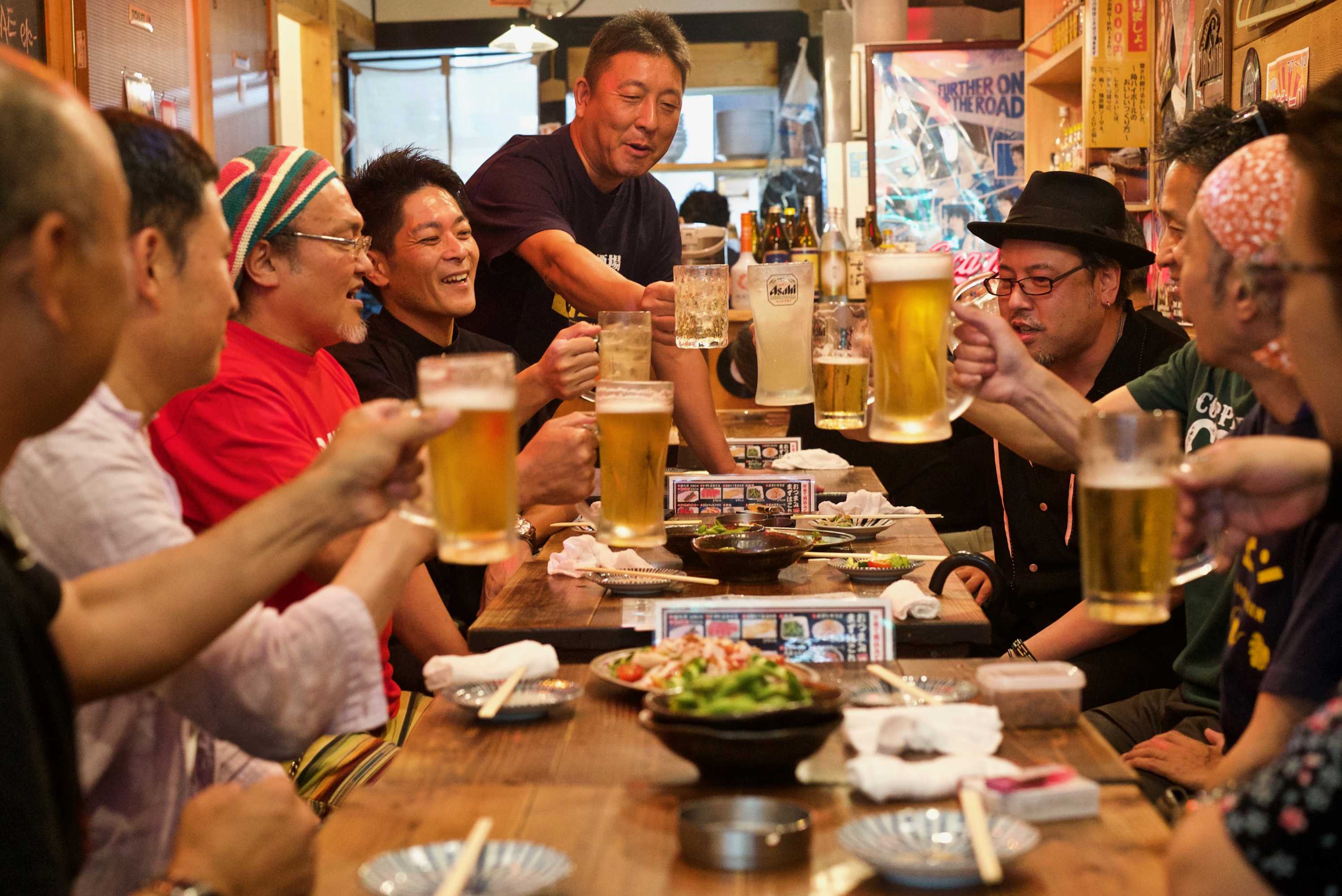 Un grupo de hombres sonrientes sentados alrededor de una mesa levantan sus vasos de cerveza a modo de brindis.