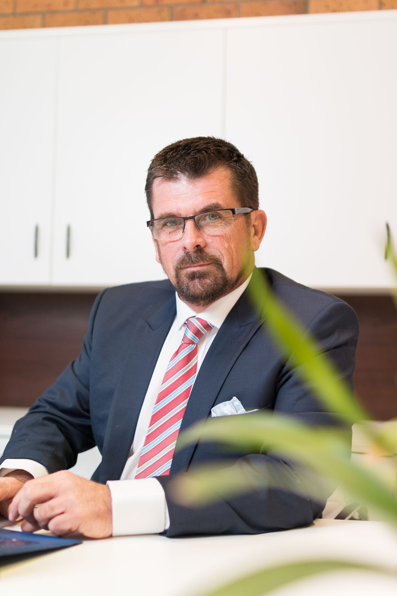 Steve Wallis is wearing a suit and tie, sitting at a desk. He is wearing glasses.