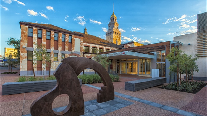 An abstract statue stands outside the stately Kalgoorlie Courthouse in front of a brilliant blue sky scattered with clouds.
