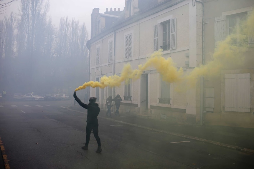 In the city of Bourges, a lone protester walks down a traditional French street with smoke grenade emitting yellow smoke.