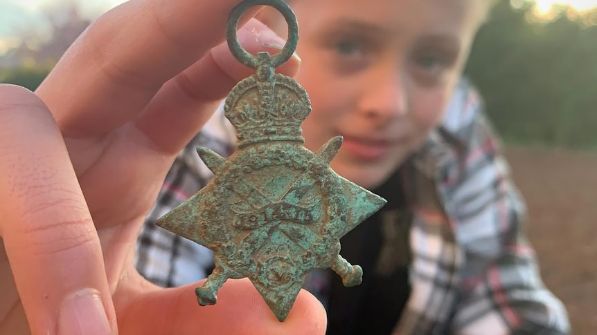 A boy of about 11 holds a war medal out in front of his face and looks at the camera.