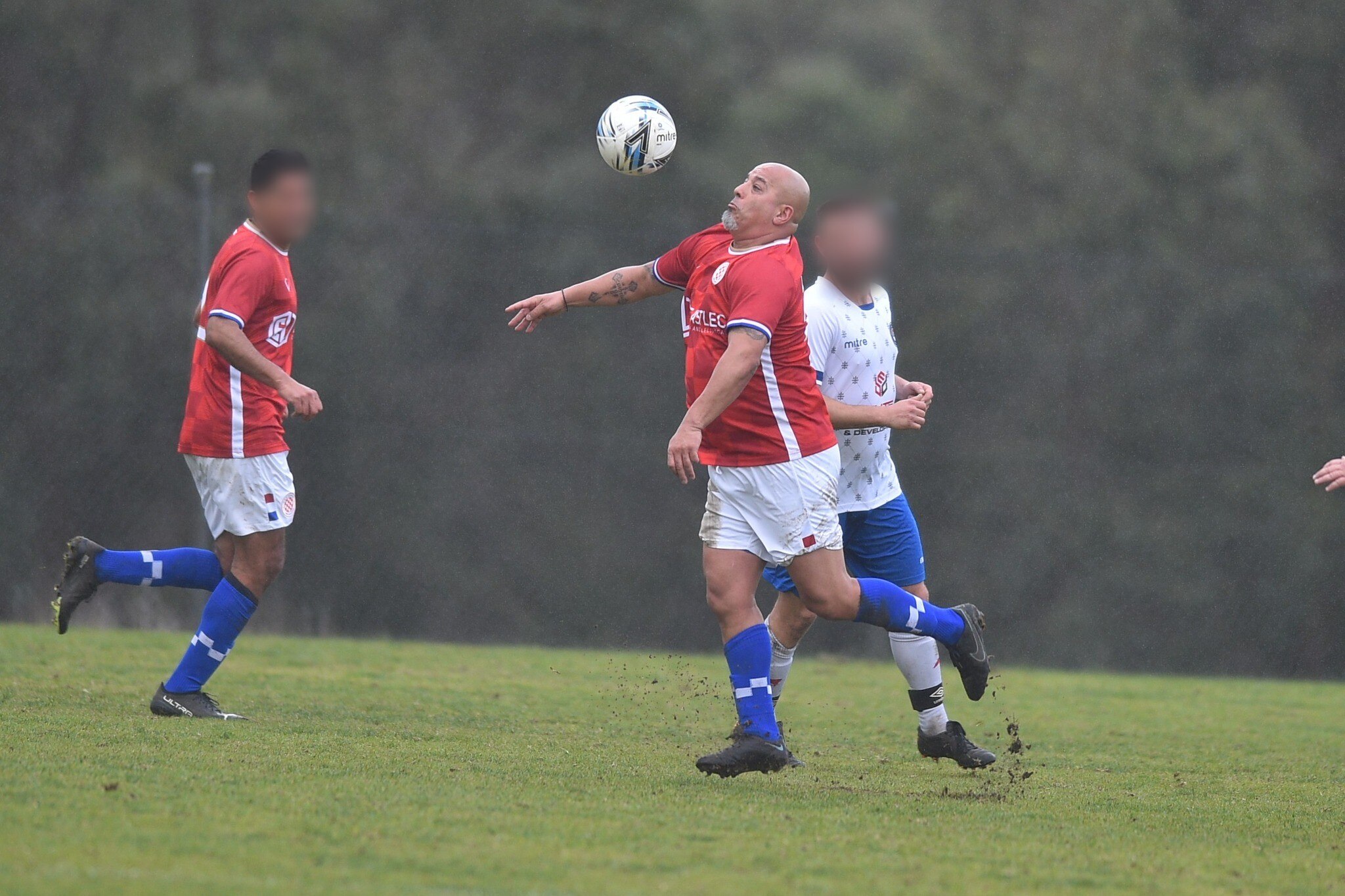 A soccer ball hangs in the air in front of a bald man in a red shirt and white shorts as two other players run behind him.
