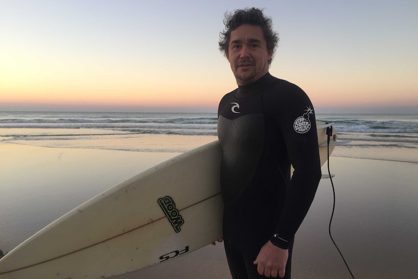 Michael Bush holds a surfboard on a beach