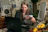 A woman speaking to students who are watching via a laptop, with an easel a still life arrangement and student artwork behind