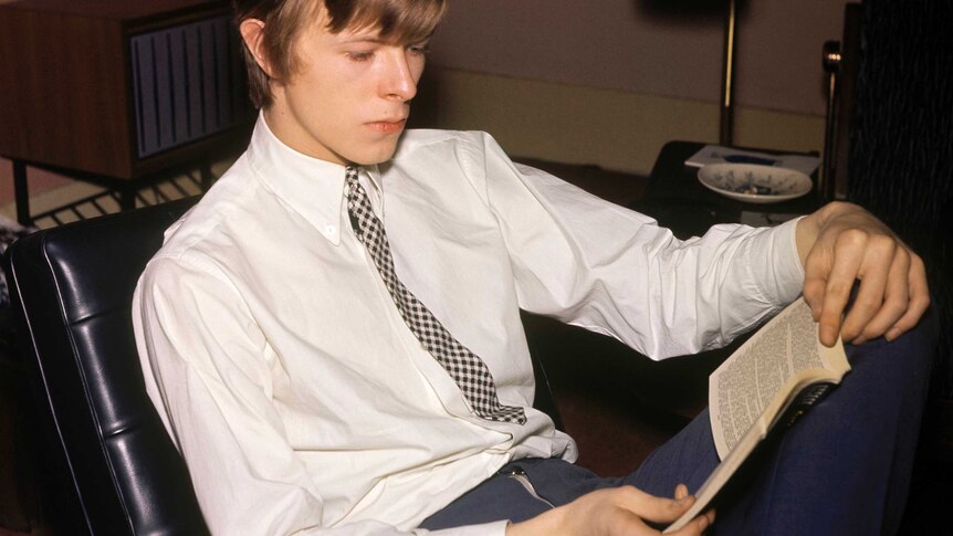 A man sitting in a hotel room wearing a white shirt and checked tie reads a novel