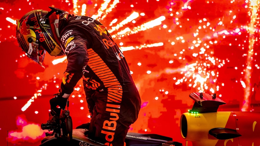 An F1 driver hops out of his car, with a firework display in the background, with red and orange lights