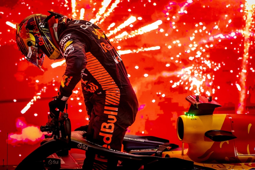 An F1 driver hops out of his car, with a firework display in the background, with red and orange lights