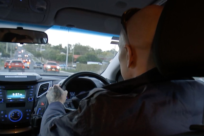 An in-car view from back seat shows a bald man driving small car on a busy city street at dusk.