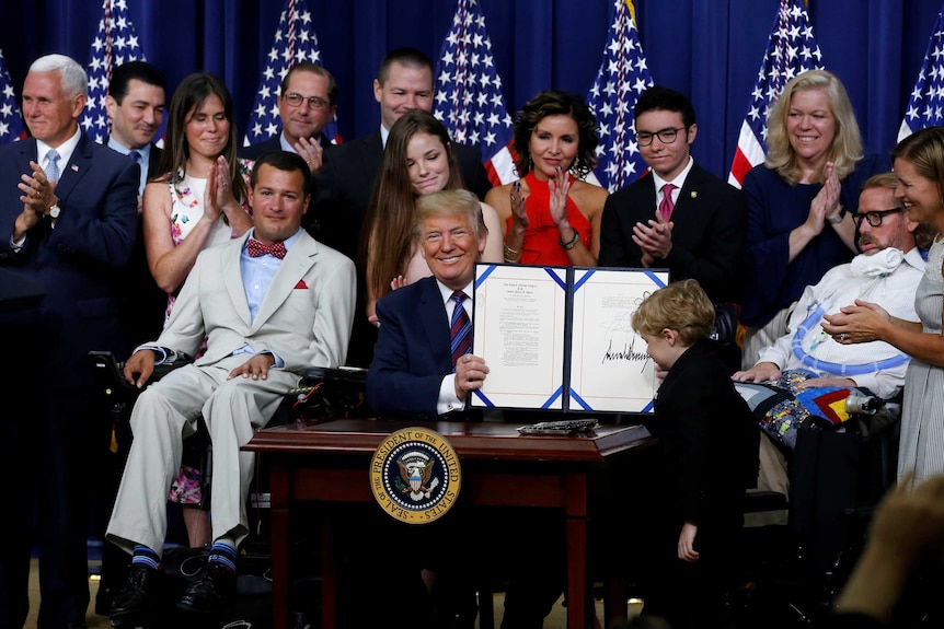Donald Trump sits at a desk holding up a signed bill surrounded by people.