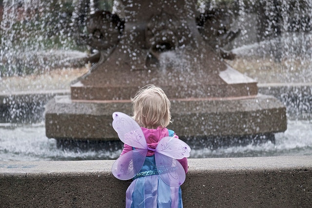 A child in a fairy costumer is on her own staring at a wishing well