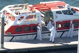 Four people in white full-body hazmat suits stand next to a white ferry-sized boat.