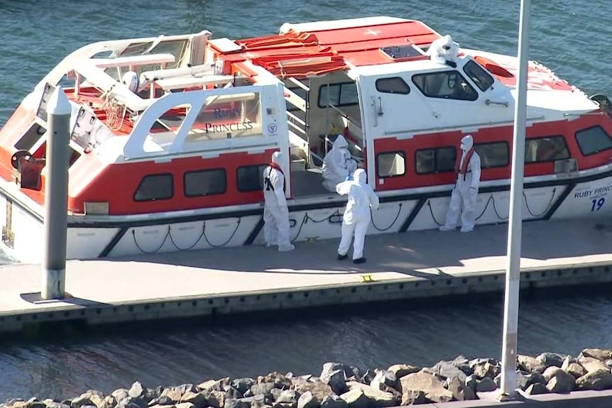 Four people in white full-body hazmat suits stand next to a white ferry-sized boat.